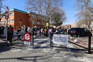 Calle urbana frente a un colegio que aparece en la imagen. Delante del paso de cebra que une tanto la acera junto al edificio como la opuesta hay dos vallas metálicas que cortan el paso. Sobre la hoja izquierda hay una señal de tráfico que prohíbe el paso de coches y motos con un panel de horarios. Sobre la hoja derecha, un cartel blanco con letras azules reza "ENTORNO ESCOLAR SEGURO Y SALUDABLE". Detrás de la valla, un gran grupo de adultos y niños cruza por un paso de peatones dirigiéndose hacia la entrada de un edificio de ladrillo naranja que tiene una colorida verja. El día es soleado y los árboles no tienen hojas.