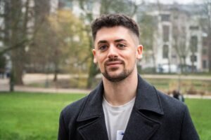 retrato fotográfico de Miguel Fuentes Sánchez, concejal de Juventud, Transición Ecológica y Movilidad Sostenible de Parla. Cabello oscuro corto. Viste un abrigo gris abierto sobre camiseta blanca. Mira a la cara con expresión amable y una leve sonrisa. El fondo es un parque urbano desenfocado con árboles, cesped y edificios.