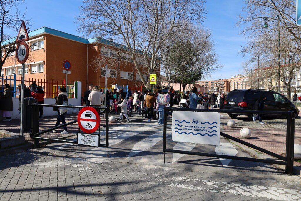 Calle urbana frente a un colegio que aparece en la imagen. Delante del paso de cebra que une tanto la acera junto al edificio como la opuesta hay dos vallas metálicas que cortan el paso. Sobre la hoja izquierda hay una señal de tráfico que prohíbe el paso de coches y motos con un panel de horarios. Sobre la hoja derecha, un cartel blanco con letras azules reza "ENTORNO ESCOLAR SEGURO Y SALUDABLE". Detrás de la valla, un gran grupo de adultos y niños cruza por un paso de peatones dirigiéndose hacia la entrada de un edificio de ladrillo naranja que tiene una colorida verja. El día es soleado y los árboles no tienen hojas.